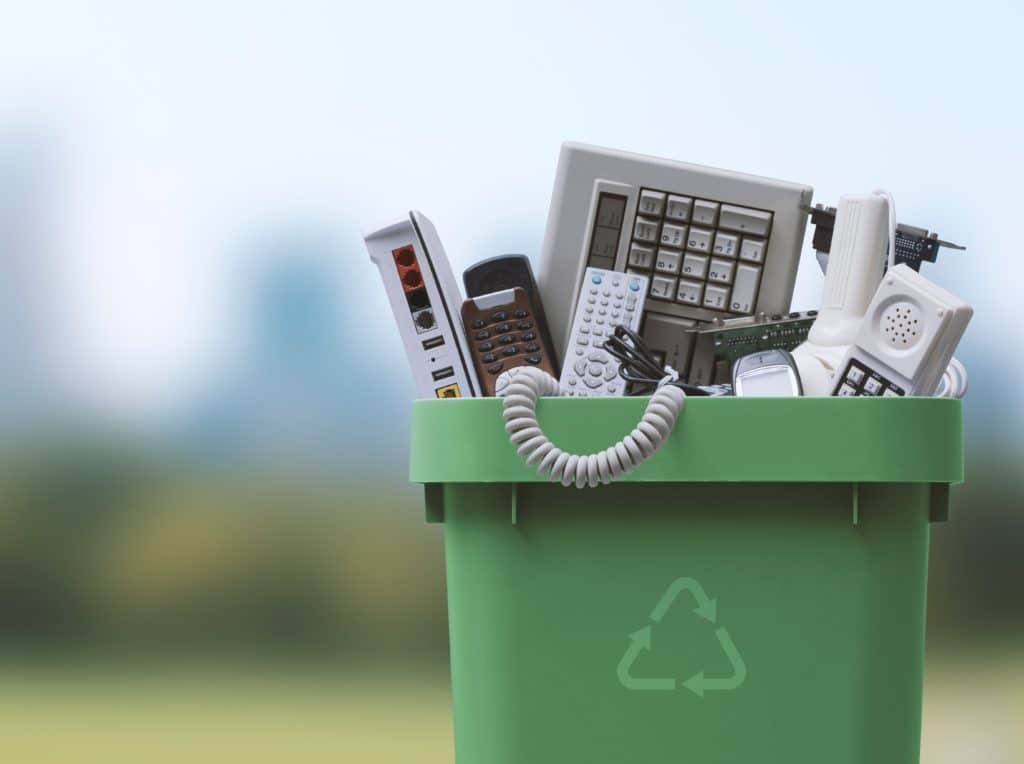 A recycling bin filled with old electronics, including phones, keyboards, computer screens, and other e-waste items.
